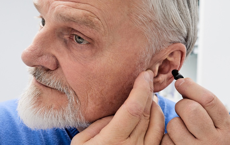 Senior man putting a hearing aid on his ear while visit his doctor at hearing clinic. Hearing solutions for elderly deafness people