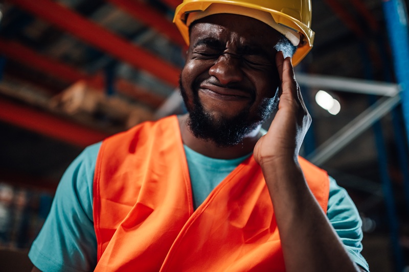 Black warehouse worker suffering from earache touching temples wearing hardhat and high visibility vest in a warehouse or storage room