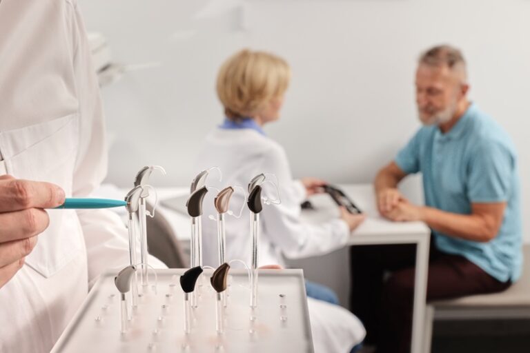 Doctor audiologist showing display stand with BTE hearing aids different types or colors while patient consultation. Hearing aids and solutions