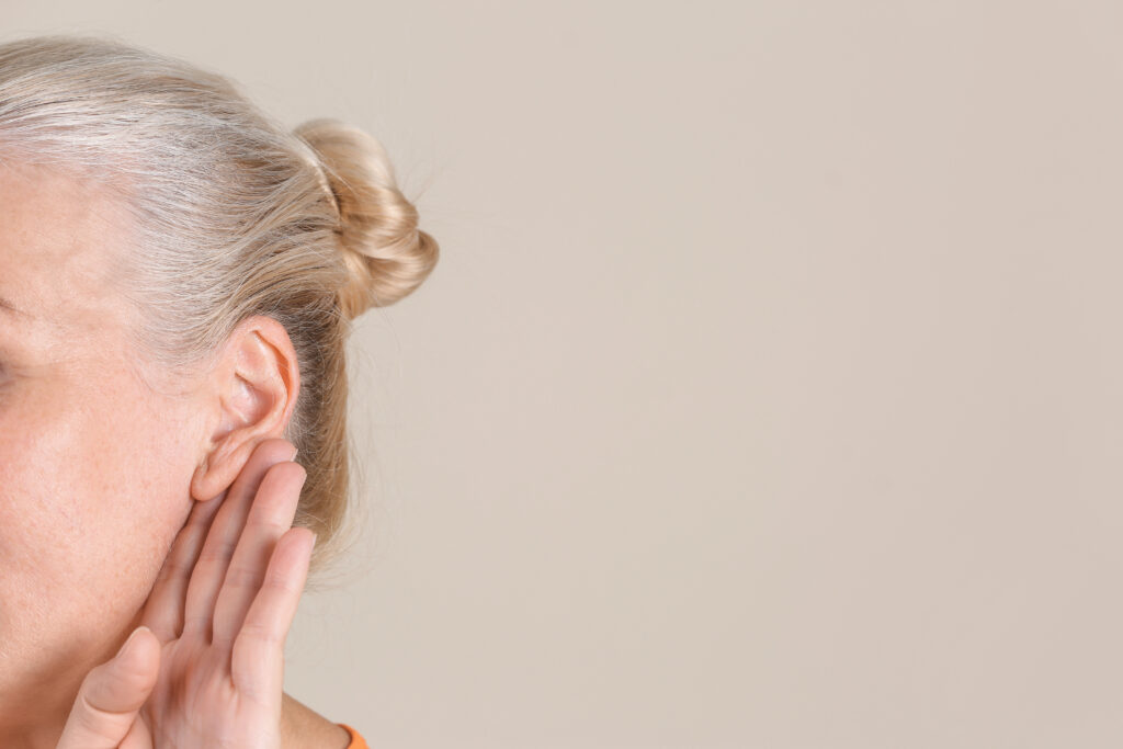 Older woman with blonde hair in a bun cupping her hand behind her ear to hear better, a common sign of hidden hearing loss